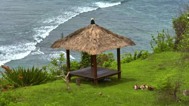 Tropical gazebo with a thatched roof providing shade on a green hillside, offering a peaceful ocean view of the serene Bali Amed coastline with a surfer in the calm water. Panoramic drone flight shot