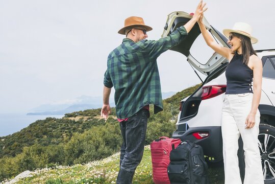 Happy Travelers tourist couple hi5 stand put luggage to white rented SUV vehicle trunk on scenic Mediterranean viewpoint. Weekend get away road trip arrival, travel preparation, planning holiday\