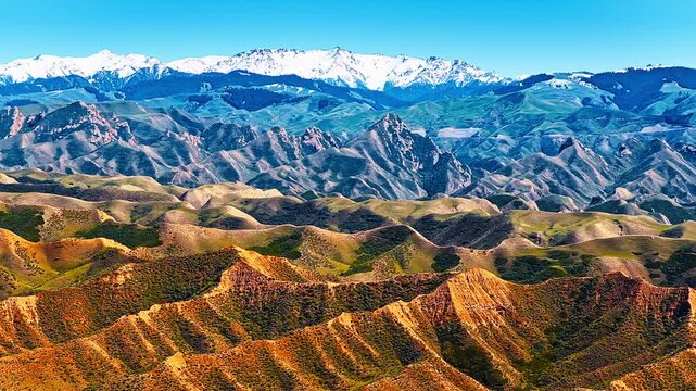 Snowy mountain peaks tower over layered colorful ridges and rolling green hills under a bright clear sky on a sunny day.