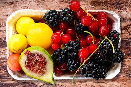 A cardboard fruit bowl containing apricots, a fig, currants, and blackberries sits on a wooden table. 