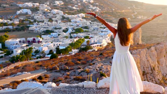 A happy woman in a white dress looks at the town of Folegandros island, Cyclades, Greece, from a viewing point during a golden summer sunset