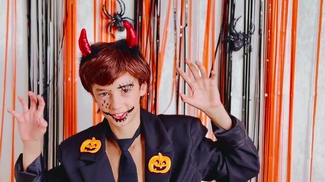 Halloween kid with devil horns headband making scary hand gestures against orange‑black streamers and spiders