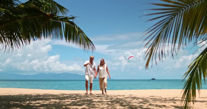 Happy mature couple holding hands and walking on sandy beach under palm trees, enjoying tropical resort vacation with ocean and blue sky in the background. Travel and relax on Samui Island, Thailand