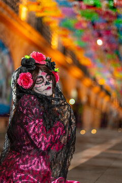 A young woman dressed as La Catrina, wearing a beautiful traditional pink Mexican dress and sugar skull makeup, praying for her deceased loved ones on the Day of the Dead beneath a canopy of *papel pi