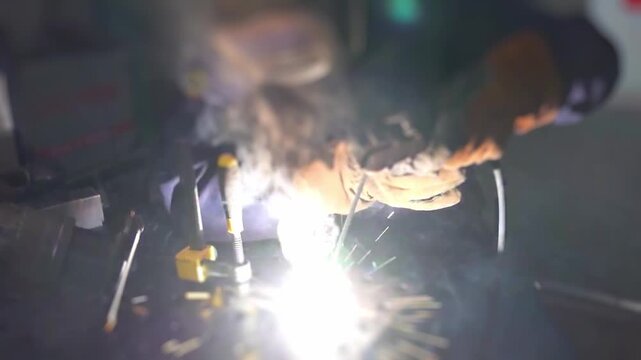 Close view of gloved hands steering welding arc onto metal workpiece, sparks burst and smoke rises as molten metal fuses during industrial fabrication work in workshop.