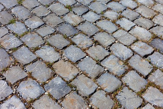 Rustic grey cobblestone pavement background texture with green grass growing between stone blocks surface