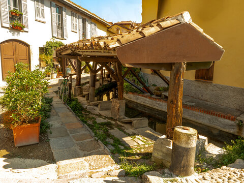 An ancient outdoor laundry basin featuring a rustic timber canopy situated along a cobblestone path in an italian town