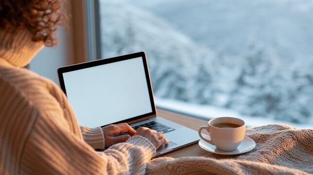 Woman using laptop beside a window with snowy outdoor view, warm sweater, coffee cup, and knitted blanket in a cosy home interior