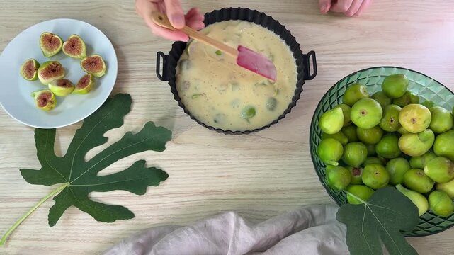 A person uses a pink spatula to stir a pale yellow batter with green fruit pieces in a black fluted baking pan on a light wood surface