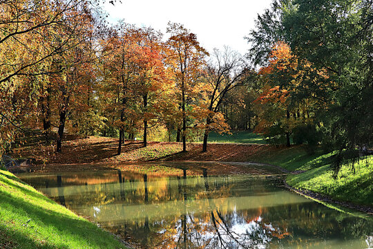 Lake surrounded by red and yellow foliage, with its reflection in the water on sunny day. Autumn park in Russia in September, picturesque autumn park landscape,vibrant autumn banner,
