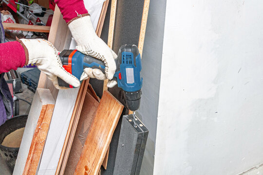 Gloved hands use a cordless drill to fasten a metal bracket beside stacked wood trim. Carpentry and home repair. DIY renovation and construction work. Workshop tools and maintenance