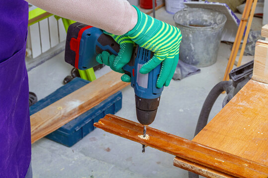 Gloved hands drilling a wooden plank with a power drill in a workshop. Woodworking and carpentry repair. DIY furniture restoration and home improvement. Practical craft and renovation