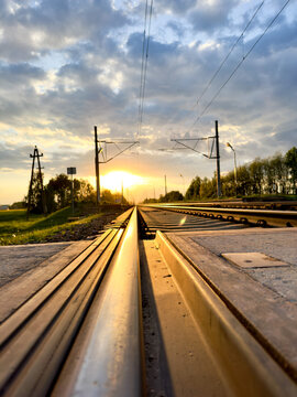 Parallel steel rails and overhead wires converge toward low golden light beneath a cloudy sky.