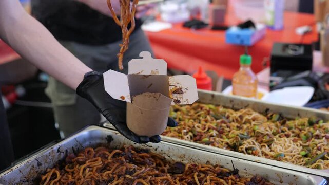 Cooked mealworms are served into cardboard cup at busy market stall. Vendor uses gloved hands and spoon to portion insect cuisine from trays of prepared food for curious customers.