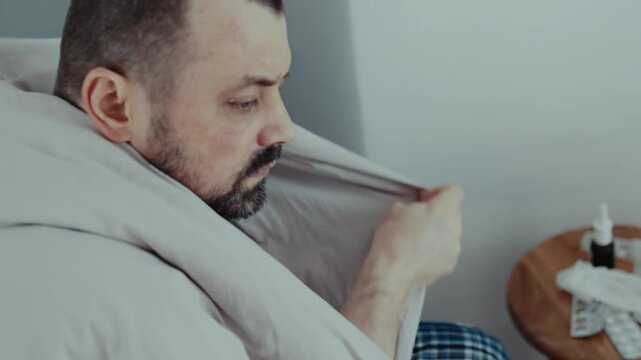 Sick man wrapped in white blanket looking down sadly in bedroom