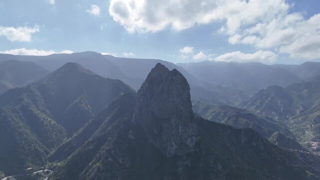 Aerial drone shot of the majestic Roque Cano mountain and surrounding rugged, lush landscape on La Gomera, Canary Islands. The drone slowly pans right, revealing mountain peaks and a distant village.