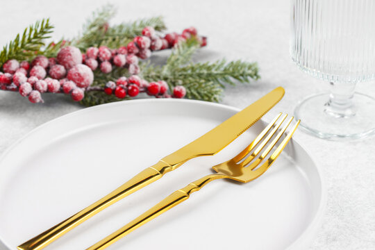 Festive holiday table setting with golden cutlery arranged on white plate with showy Christmas tree branch with red holly berry on light gray textured background. Knife and fork close up, macro