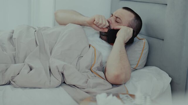 Sick man lying under blanket in bed coughing into hand