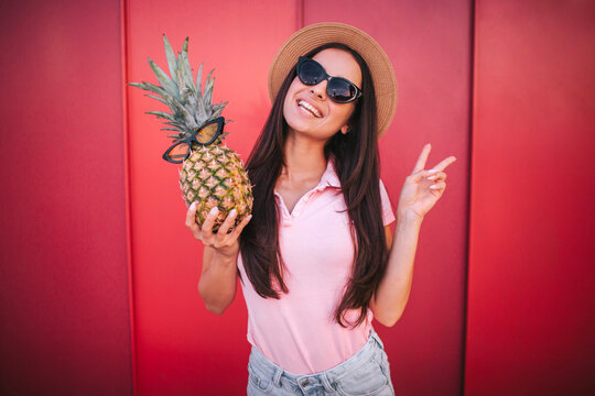 Beautiful woman stands and poses on camera. She shows the piece symbol with her fingers. Woman has pineapple in right hand. Both she and the fruit have glasses. Isolatd on red and striped background.