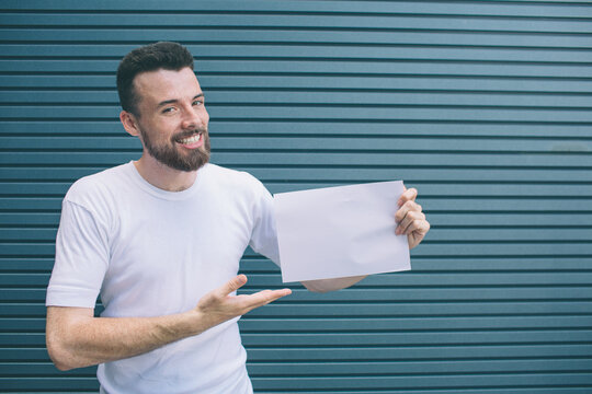 Nice and positive man iss standing and smiling. He is posing and looking at camera. Guy is holding piece of paper and poiting on it with hand. Isolated on striped and blue background.