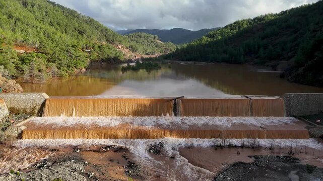 Namnam River in Koycegiz, Mugla, Turkey, with muddy floodwater pouring over a concrete stepped weir, a brown reservoir and pine-forested hills behind. Aerial drone view