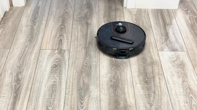 Modern black robotic vacuum cleaner cleaning a light wooden laminate floor indoors. Smart wireless home appliance sweeping dust in a cozy living room or apartment house. Domestic automatic hygiene. 