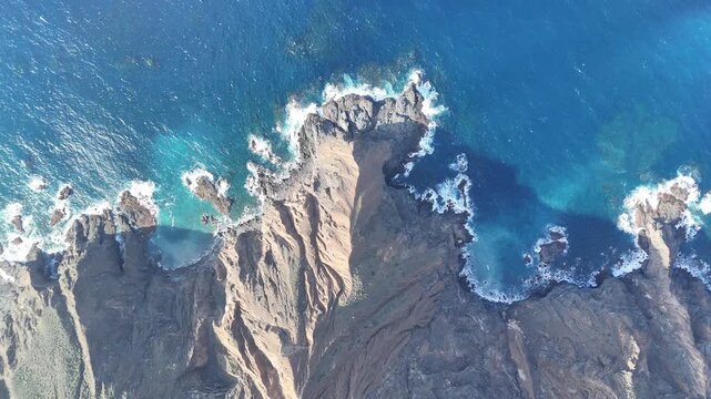 A slow aerial drone shot pans right over the dramatic, rugged coastline of La Gomera, Canary Islands. The deep blue ocean meets rocky cliffs, with waves breaking into white foam.