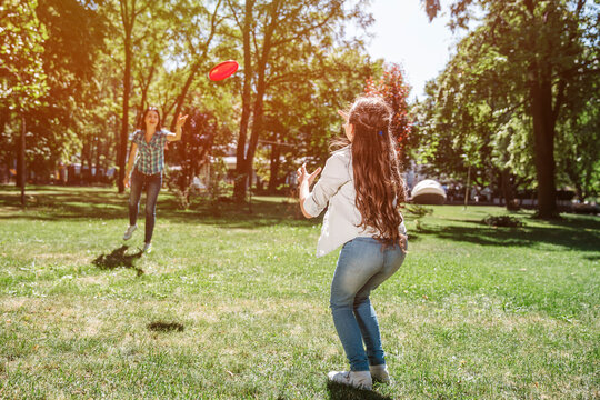 Mom and child are standing in front of ech other and playing with frisbee. Woman is throwing it to girl. Child is planning to catch it. Frisbee has red color.