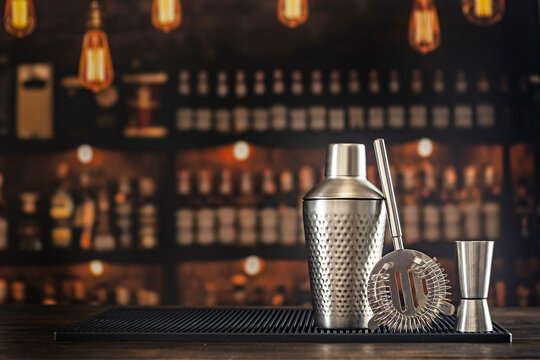 Cocktail shaker strainer jigger on black rubber mat. Blurred bar shelves bottles warm lamps background. Metal barware tools for drink preparation.