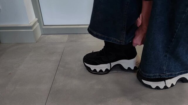 Woman puts on a second black sneaker, first attempting to do so while balancing before placing her foot on the floor, everyday home routine, horizontal video.