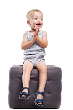 Child sits on a gray ottoman and claps hands while smiling in a studio setting
