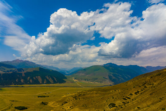 Wide shot of a mountain valley featuring rolling green hills and distant layered mountains. A winding path leads through the verdant valley floor, with sparse vegetation on the dry hillside.