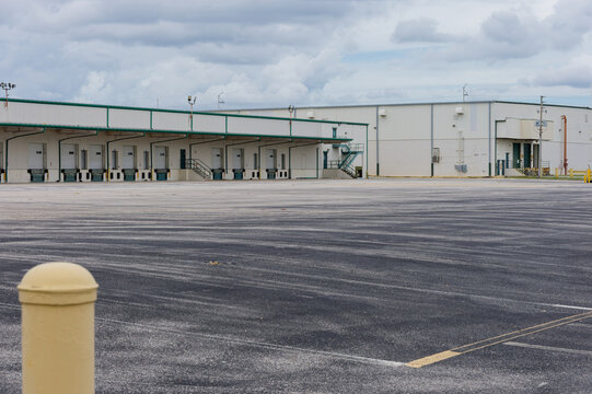 Empty warehouse loading docks and a large asphalt yard at an industrial distribution center under a cloudy sky, featuring closed bay doors, a yellow bollard and utilitarian architecture for logistics 