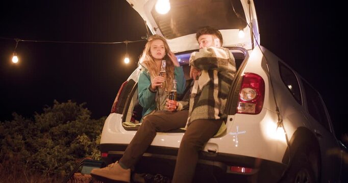 Friends enjoy snacks and drinks together at night by the car in a remote area with string lights in the background