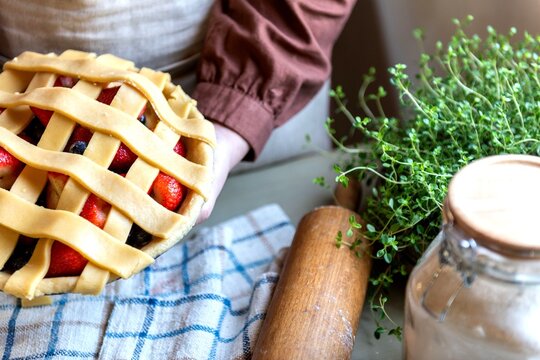 Person holding a lattice-topped berry pie beside fresh herbs and a rolling pin.