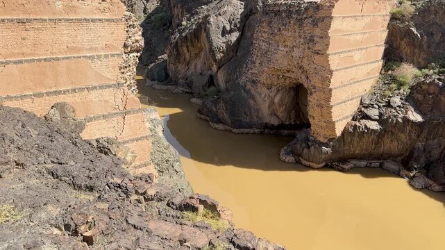 Ancient dam ruins and a muddy river flowing through a rocky gorge in Saudi Arabia.