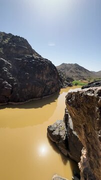 Muddy wadi flows through a rocky canyon past ancient stepped brick dam ruins.