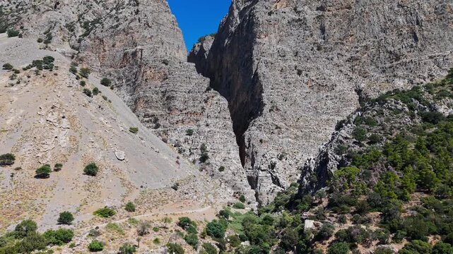 Narrow entrance to Ha Gorge in eastern Crete, massive sheer limestone cliffs towering over scree slopes and green Mediterranean scrub, aerial view