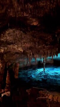 Panning shot of illuminated stalactites and stalagmites in underground cave with water