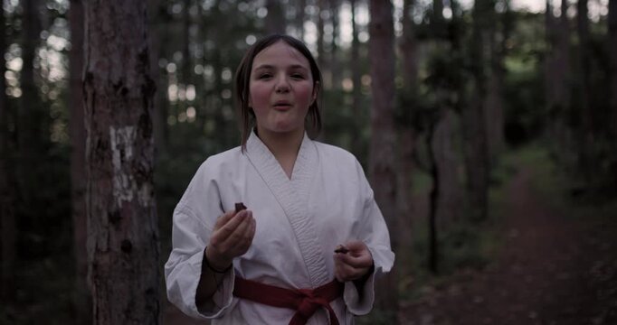A young girl in a white karate gi and red belt enjoys a peaceful snack in the serene forest before starting her martial arts training on a blue mat, embracing nature's calm.