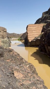 Muddy wadi water flowing past ancient dam ruins in a rocky Saudi Arabian canyon.