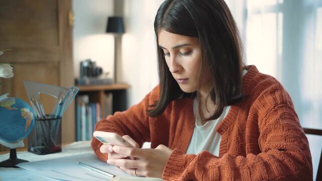 Young Latina student using her smartphone instead of studying, sitting at her desk with her academic work set aside. Concept of student life, digital distraction, procrastination, social media, mobile