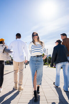 Vertical. Young woman walks toward the camera while friends continue ahead on a sunny promenade, creating a lively urban lifestyle and travel scene blue sky.