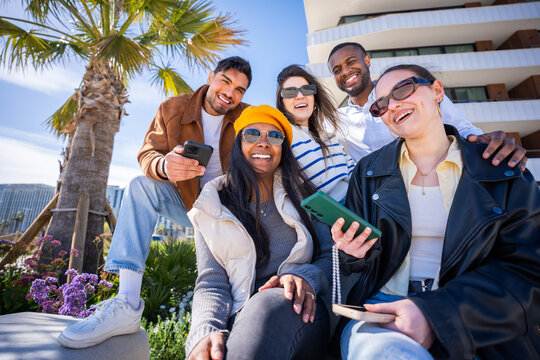 Five multicultural friends sit together and smile with phones in hand, enjoying leisure time and digital connection in a sunny residential district.