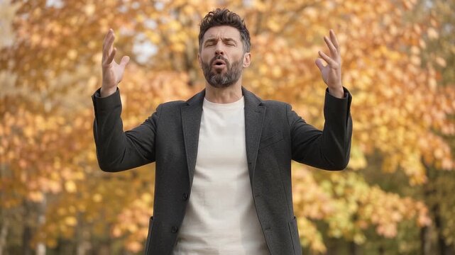 Bearded man in a charcoal jacket raises both open hands beside his head, looks upward and opens his mouth before lowering his arms, standing against bright golden autumn foliage in a park.
