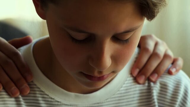 Soft light falls on a young boy's face. His head is bowed, eyes lowered. Two hands rest gently on his shoulders. He wears a simple striped shirt. The moment feels quiet and comforting