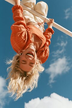 Upside down woman laughing while gripping outdoor bar, windblown hair and bright blue sky, park
