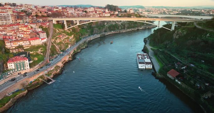 Aerial view drone flight over historic Porto cityscape along the Douro River, the Infante Dom Henrique bridge, a river cruise boat, and surrounding urban architecture at sunset. Travel Porto, Portugal