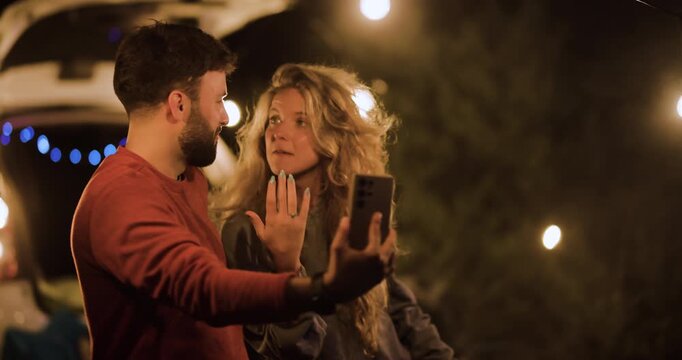 Couple enjoying night under string lights while taking selfies at a campsite, sharing laughs and smiles by their car with glowing decorations in the background