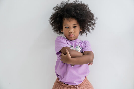 Portrait of black African American toddler girl in the bad mood. Angry kid arms fold while standing over white wall. Burnout kindergarten girl feeling sad.